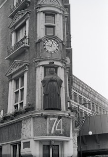 A historic building facade featuring intricate stonework and a prominent clock with Roman numerals. Below the clock is a statue of a figure in robes, possibly a monk or scholar. The address number 174 is prominently displayed in mosaic tiles. Windows with ornate railings and decorative architectural elements adorn the structure.