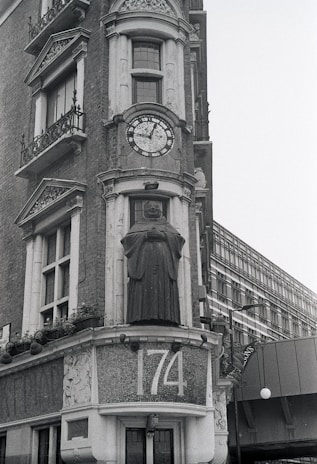 A historic building facade featuring intricate stonework and a prominent clock with Roman numerals. Below the clock is a statue of a figure in robes, possibly a monk or scholar. The address number 174 is prominently displayed in mosaic tiles. Windows with ornate railings and decorative architectural elements adorn the structure.