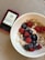 A white bowl filled with oatmeal topped with fresh raspberries, blueberries, and a sprinkle of granola. A spoon is resting inside the bowl. Next to the bowl, there is a digital device displaying a numerical reading, possibly a glucose meter, placed on a woven placemat.