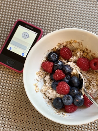 A white bowl filled with oatmeal topped with fresh raspberries, blueberries, and a sprinkle of granola. A spoon is resting inside the bowl. Next to the bowl, there is a digital device displaying a numerical reading, possibly a glucose meter, placed on a woven placemat.