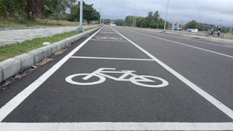 A newly paved road with designated bicycle lanes marked by white bike symbols. The road is flanked by concrete pavements and grassy areas. Sparse streetlights are visible along the path, and trees can be seen in the background under an overcast sky.