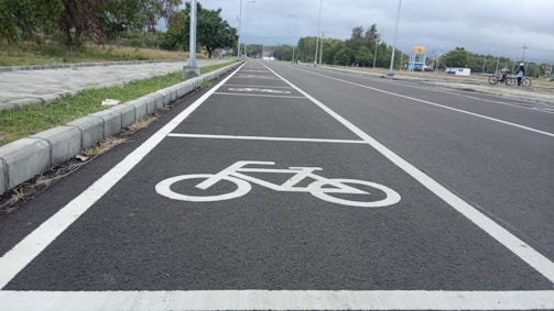 A newly paved road with designated bicycle lanes marked by white bike symbols. The road is flanked by concrete pavements and grassy areas. Sparse streetlights are visible along the path, and trees can be seen in the background under an overcast sky.