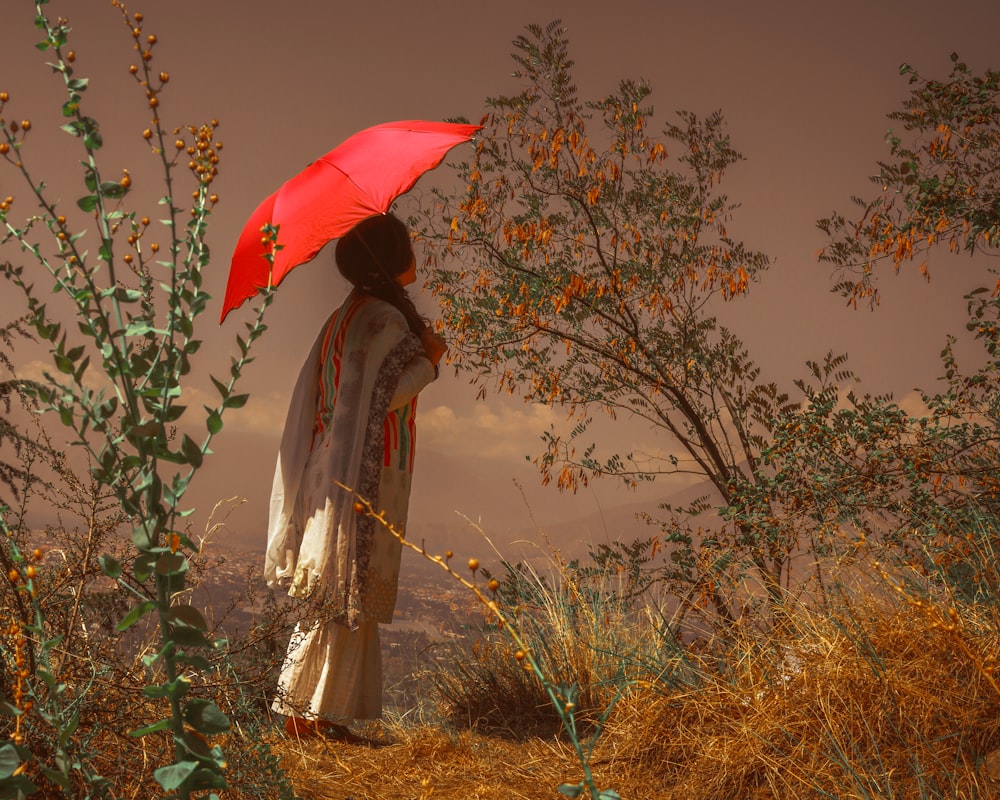 Mujer en vestido blanco y marrón sosteniendo paraguas rojo de pie en el campo de hierba verde durante el día