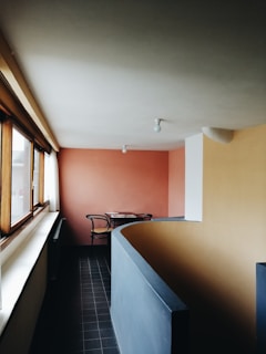 A modern interior space with clean lines and minimalist design. The room features a pink accent wall and a contrasting tan wall, with a window running along one side. Natural light illuminates the space, highlighting the curved, blue-gray partition. In the corner, there is a small table with two black chairs.