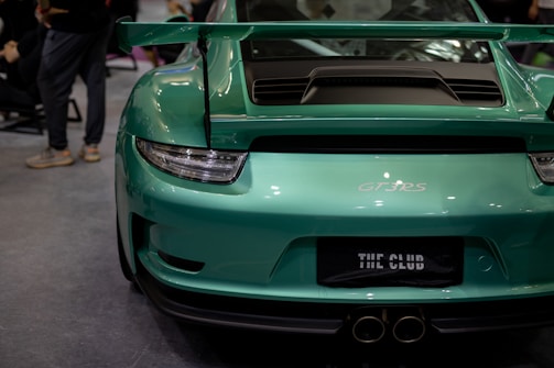 A vibrant green sports car is parked indoors, displaying a sleek design with a prominent rear wing and dual exhaust pipes. The car is a GT3 RS model, and the license plate displays the words 'THE CLUB'. Several people are visible in the background, suggesting an indoor event or showroom environment.