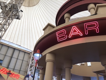 An interior view of a building, featuring a domed ceiling with a central structure and industrial elements. A neon sign displaying the word 'BAR' is prominently visible on a circular architectural feature. The scene includes signs for 'Hotshots' and 'Costa', adding a commercial vibe to the setting.