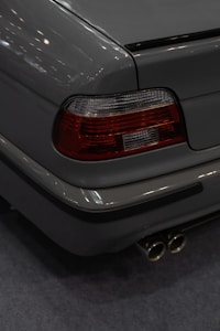 Rear view of a car, focusing on the tail light and exhaust pipes. The tail light is red with white sections, and the car body is a glossy gray. The dual exhaust pipes are stainless steel.