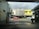 A street scene from inside a parking area, looking out towards a row of small business establishments. The buildings feature varied architecture, one in yellow and others in neutral colors. A red car and a white car are visible on the road. There is a visible 'For Lease' sign and storefronts for Sushi Metro and Cordelia's Cafe.