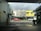 A street scene from inside a parking area, looking out towards a row of small business establishments. The buildings feature varied architecture, one in yellow and others in neutral colors. A red car and a white car are visible on the road. There is a visible 'For Lease' sign and storefronts for Sushi Metro and Cordelia's Cafe.