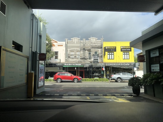 A street scene from inside a parking area, looking out towards a row of small business establishments. The buildings feature varied architecture, one in yellow and others in neutral colors. A red car and a white car are visible on the road. There is a visible 'For Lease' sign and storefronts for Sushi Metro and Cordelia's Cafe.