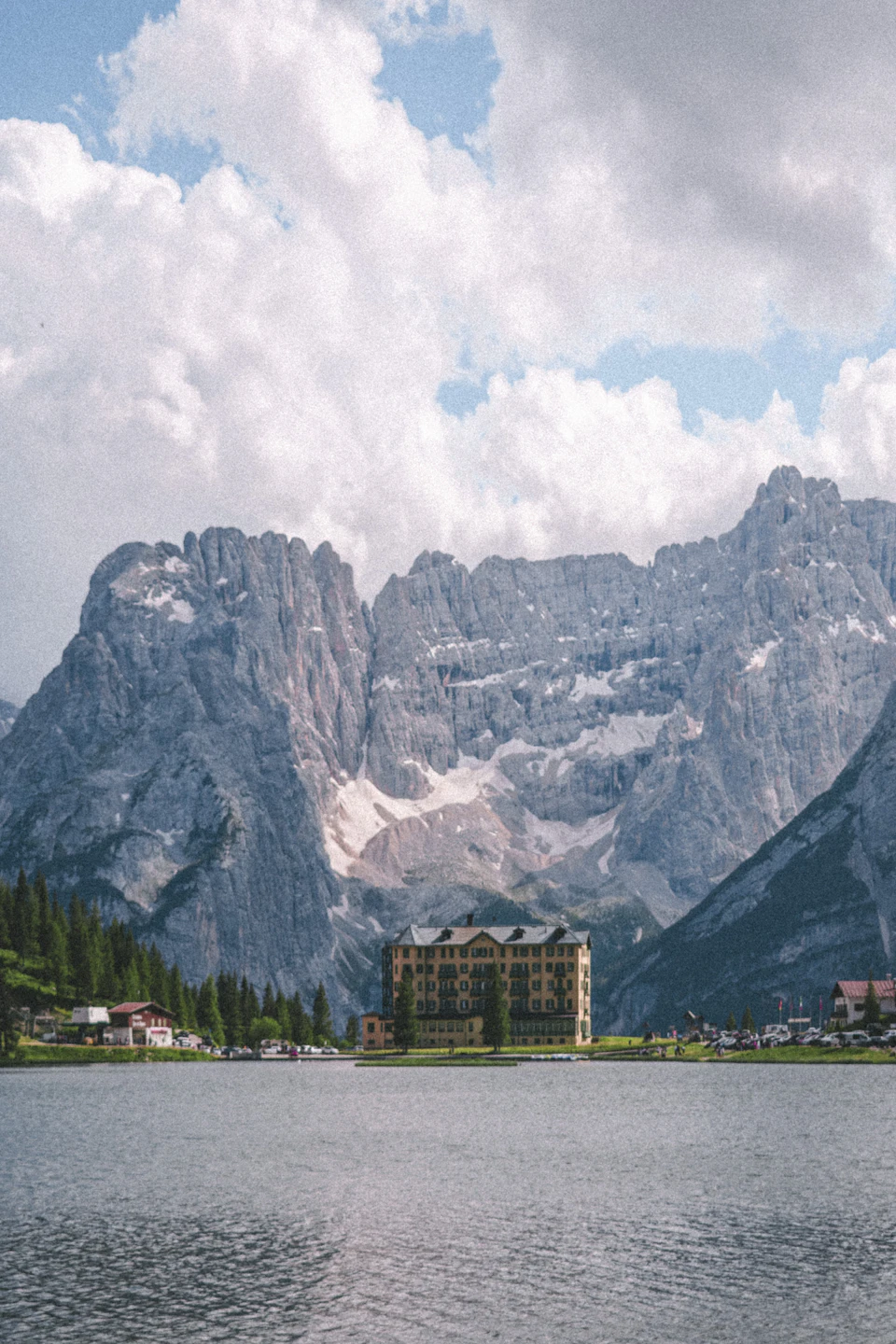 The image depicts a serene mountain lake surrounded by snow-capped mountains and buildings on its banks. The calm, light blue water reflects the clear sky above, creating a picturesque scene. On one side of the lake, there is a large building with a brown roof, possibly an old hotel or resort. The other side features several smaller structures, suggesting that this location may be a popular tourist destination.