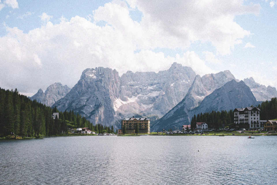 The image depicts a serene mountain lake surrounded by lush green trees and majestic mountains with snow-capped peaks. The calm water reflects the clear blue sky, creating a beautiful mirror image. On one side of the lake, there is a large building with a yellow roof, possibly a hotel or resort, nestled among the trees. In the distance, several other buildings can be seen, suggesting that this location may be a popular tourist destination.