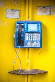 A blue public payphone mounted on a bright yellow wall, showing signs of wear and rust. The handset is black, and the phone has a keypad with multiple buttons. There is a metallic cord connecting the handset to the phone. Notices are posted above the phone, indicating it is for international calls.