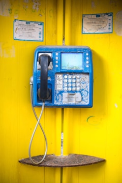 A blue public payphone mounted on a bright yellow wall, showing signs of wear and rust. The handset is black, and the phone has a keypad with multiple buttons. There is a metallic cord connecting the handset to the phone. Notices are posted above the phone, indicating it is for international calls.