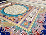 An ornate and intricate piece of decorative art featuring vibrant colors and traditional Islamic calligraphy. The design includes geometric patterns and floral motifs, with script inscribed within a central circle surrounded by rich, swirling details.