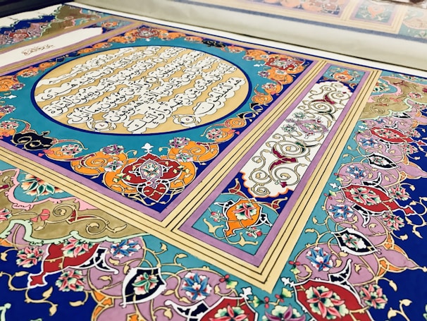 An ornate and intricate piece of decorative art featuring vibrant colors and traditional Islamic calligraphy. The design includes geometric patterns and floral motifs, with script inscribed within a central circle surrounded by rich, swirling details.