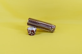 A pack of stacked plastic cups wrapped in transparent plastic lies horizontally on a plain yellow surface. Next to it is an ornate brown and white paper cup with a floral pattern, standing upright.