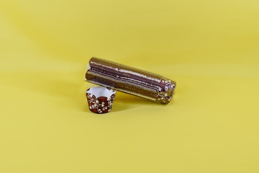 A pack of stacked plastic cups wrapped in transparent plastic lies horizontally on a plain yellow surface. Next to it is an ornate brown and white paper cup with a floral pattern, standing upright.