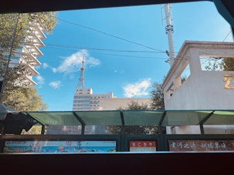 A cityscape featuring a tall communication tower and several buildings under a bright blue sky with scattered clouds. Trees frame parts of the scene, providing a touch of greenery. In the foreground, there is a partially transparent overhang, possibly a bus stop, with signage displaying advertisements and text.