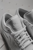 A close-up view of white sneakers displaying intricate details including laces, stitching, and logos on the tongue and side. The background includes small, repeated numbers '23'. The sneakers have a modern and sporty design.