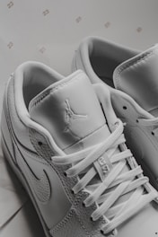 A close-up view of white sneakers displaying intricate details including laces, stitching, and logos on the tongue and side. The background includes small, repeated numbers '23'. The sneakers have a modern and sporty design.