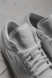 A close-up view of white sneakers displaying intricate details including laces, stitching, and logos on the tongue and side. The background includes small, repeated numbers '23'. The sneakers have a modern and sporty design.