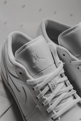 A close-up view of white sneakers displaying intricate details including laces, stitching, and logos on the tongue and side. The background includes small, repeated numbers '23'. The sneakers have a modern and sporty design.