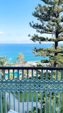 A picturesque view from a balcony over a lush garden with palm trees, leading to a turquoise sea under a clear blue sky. A decorative white railing is in the foreground. The garden features well-maintained greenery and a path, with sunbathers visible on the beach in the distance.