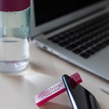 A water bottle with a magenta lid sits on a desk next to a laptop. A small pink object with the text 'quokka' is positioned near a smartphone, which is slightly tipped up.