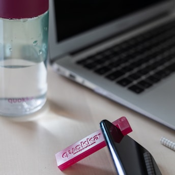 A water bottle with a magenta lid sits on a desk next to a laptop. A small pink object with the text 'quokka' is positioned near a smartphone, which is slightly tipped up.