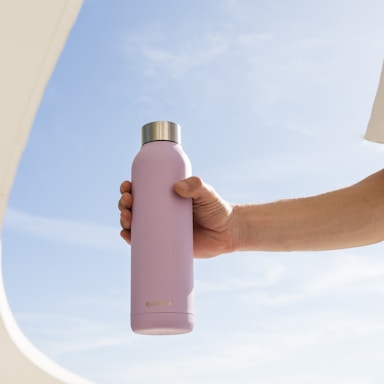 A hand holds a pastel-colored insulated water bottle against a clear blue sky. The bottle has a metallic cap and is branded with the name 'Quokka' near the bottom.