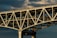 A large, sturdy bridge with complex lattice work and structural beams, showcasing its engineering design. The bridge has an industrial look, with overhead clouds creating a dramatic background.