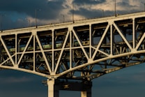 A large, sturdy bridge with complex lattice work and structural beams, showcasing its engineering design. The bridge has an industrial look, with overhead clouds creating a dramatic background.
