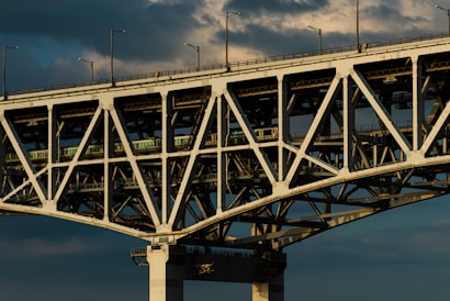 A large, sturdy bridge with complex lattice work and structural beams, showcasing its engineering design. The bridge has an industrial look, with overhead clouds creating a dramatic background.