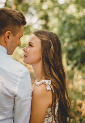 A young couple stands closely facing each other in a natural outdoor setting with lush green foliage blurred in the background. The woman gazes upward towards the man's face with a gentle, serene expression. Her long hair cascades down her back, and she wears a floral dress with tied straps.