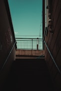 Concrete stairs lead upwards towards a metal fence, silhouetted against a teal sky. The scene features urban elements such as pipes and graffiti on the walls, suggesting an industrial or city environment.