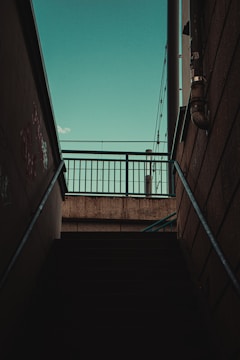 Concrete stairs lead upwards towards a metal fence, silhouetted against a teal sky. The scene features urban elements such as pipes and graffiti on the walls, suggesting an industrial or city environment.