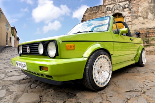 A vibrant green convertible car is parked on a cobblestone street in front of a historic stone building. The car features distinct circular headlights and alloy wheels, reflecting modern design elements against the rustic background. The sky is partly cloudy, adding contrast to the scene.