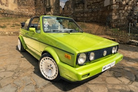 A lime green vintage convertible car with a black soft top is parked on cobblestone pavement. The car features distinctive retro headlights and a front bumper with a license plate displaying a club logo. In the background, there's a stone building with an old, textured wall, adding a historic touch to the setting.