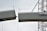 A construction worker is positioned between two large steel beams, with safety cables attached. The scene appears to be part of a bridge or building under construction, supported by a crane. The sky is overcast, providing a neutral background.