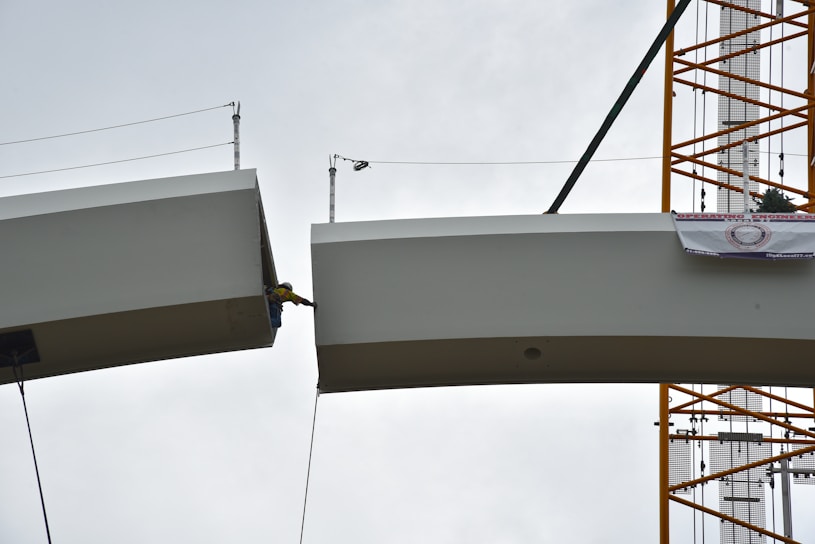A construction worker is positioned between two large steel beams, with safety cables attached. The scene appears to be part of a bridge or building under construction, supported by a crane. The sky is overcast, providing a neutral background.