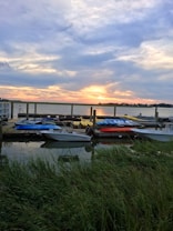 The image captures a picturesque sunset over a lake with a wooden dock in the foreground. Several kayaks and small boats are lined up on the dock. The sky is filled with dramatic clouds painted in hues of orange and blue, reflecting on the water below. Tall grass lines the shores, adding a touch of nature to the serene scene.