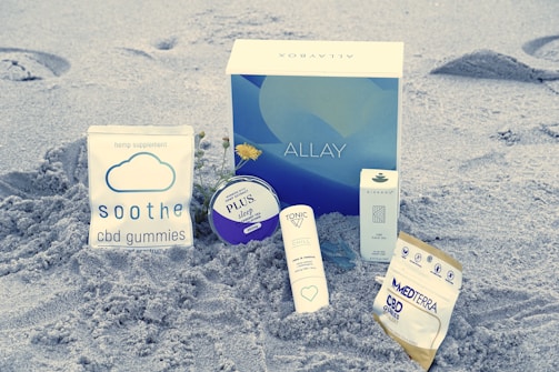 Products related to CBD are placed on the sand. Items include soothe cbd gummies, a blue and white box labeled ALLAY, a small container labeled PLUS sleep, a white box labeled OLEO CBD face oil, and packages labeled TONIC and MEDTERRA.