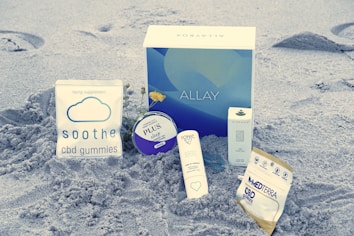 Products related to CBD are placed on the sand. Items include soothe cbd gummies, a blue and white box labeled ALLAY, a small container labeled PLUS sleep, a white box labeled OLEO CBD face oil, and packages labeled TONIC and MEDTERRA.