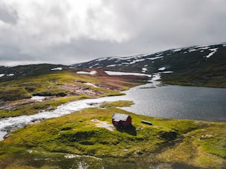 A small red cabin is situated on lush green terrain near a flowing river and adjacent to a lake. Snow-specked hills rise in the background under an overcast sky, casting a serene and isolated atmosphere.