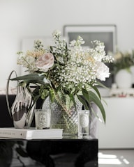 A bouquet of flowers, including roses and baby's breath, is arranged in a textured glass vase. The setting appears to be a modern interior, with a metallic jug, scented candle, and a book titled 'The Kinfolk Home' on the table. Two bottles labeled 'CYPRES' and 'FIGUIER' are included in the arrangement, suggesting a fragrant or decorative theme.