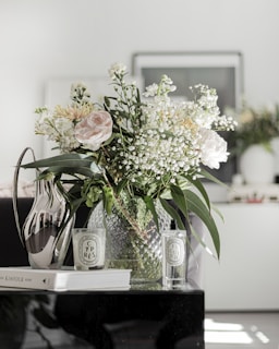 A bouquet of flowers, including roses and baby's breath, is arranged in a textured glass vase. The setting appears to be a modern interior, with a metallic jug, scented candle, and a book titled 'The Kinfolk Home' on the table. Two bottles labeled 'CYPRES' and 'FIGUIER' are included in the arrangement, suggesting a fragrant or decorative theme.