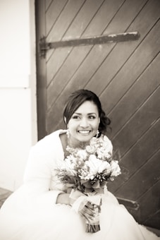 A woman dressed in a wedding gown is sitting in front of a wooden door, holding a bouquet of flowers and smiling brightly. Her hair is styled elegantly, and she wears sparkling earrings. The setting appears rustic, adding charm to the scene.