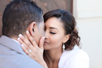 A couple is sharing an intimate moment with their faces close together. The woman is wearing elegant earrings and a white garment, and she gently holds the man's face with her hand. The background is softly blurred.