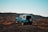 A small off-road vehicle is perched atop a grassy hill, with expansive mountain ranges in the background under a clear blue sky. The vehicle is painted in a vibrant blue with a white roof and appears rugged, suggesting it's suited for adventure.